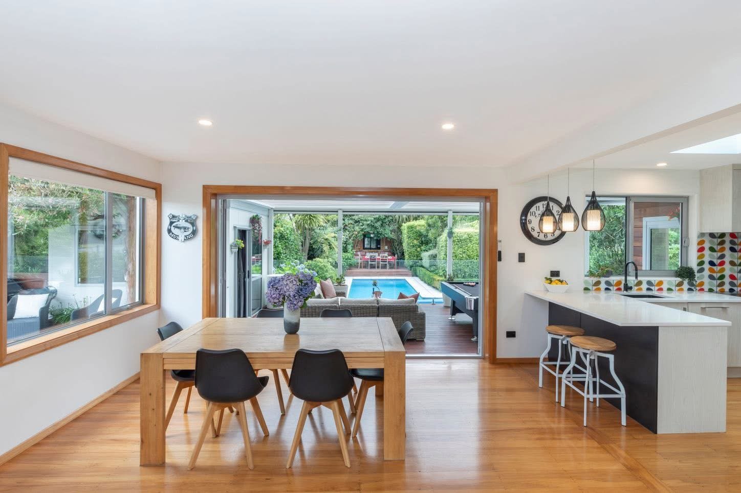 Modern open-plan home interior with wooden dining table, black chairs, kitchen area, and view of backyard pool through large glass doors